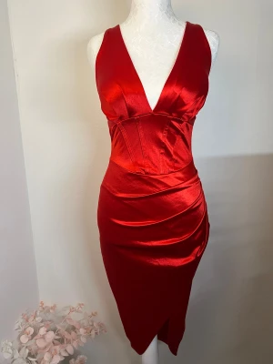 Klänning - Snygg röd klänning i satin med djup v-ringning och breda axelband samt inbyggda kupor. Figurnära passform och omlott-detalj framtill som skapar en slits. Använd en gång, i utmärkt skick ❤️