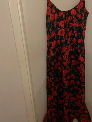 Långklänning med röda rosor - Svart långklänning med röda rosor och gröna blad i ett blommigt mönster. Klänningen har smala axelband och en lätt utställd nederdel. Perfekt för dig som vill sticka ut med en romantisk och färgstark look.