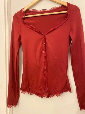 Röd blus med spets och knappar - Säljer en röd blus med lång ärm och v-ringad hals. Blusen har fina spetsdetaljer längs halsringningen, knäppningen och nederkanten. Knappar framtill och mjukt, stretchigt material som sitter snyggt på kroppen. Perfekt för dig som gillar romantiska detaljer.
