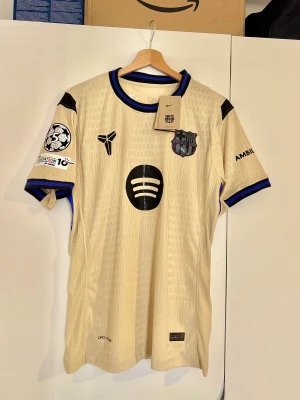 Beige FC Barcelona 25/26 Raphinha Nike tröja - Säljer en beige FC Barcelona fotbollströja med svarta och blå detaljer, Raphinha 11 på ryggen och Spotify-logga på bröstet. Märkt med Nike, FCB-emblem och Champions League-märke på ärmen. Tillverkad i lätt, ventilerande polyester med subtilt mönster och ribbad krage.