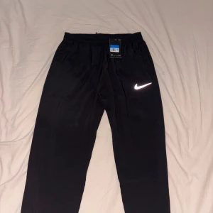 Svarta träningsbyxor från Nike - Svarta träningsbyxor från Nike med elastisk midja och klassisk vit swoosh-logga på låret. Byxorna har en rak passform och är tillverkade i ett lätt och bekvämt syntetmaterial som passar perfekt för sport och träning.