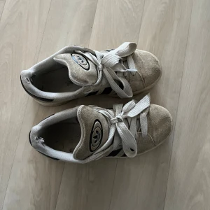 Adidas Campus sneakers beige/svart - Säljer ett par Adidas Campus sneakers i beige mocka med svarta ränder och gummisula. Klassisk modell med snörning och rund tå. Skorna har Adidas logga på plösen och sidan. Perfekta för dig som gillar retrostil och streetwear.