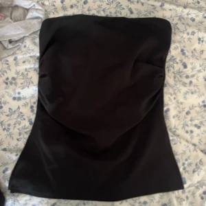 Svart axelbandslös tubtopp - Säljer en stilren svart tubtopp med axelbandslös design. Toppen har en lätt figurnära passform och slät yta, perfekt att styla till olika looks. Materialet känns mjukt och stretchigt, vilket gör den bekväm att bära. Enkel och clean look som funkar till mycket.