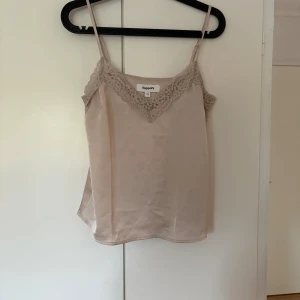 Beige linnetopp med spets från Kappahl - Supersöt beige linnetopp från Kappahl i storlek XS. Toppen har tunna axelband och v-ringning med fina spetsdetaljer längs kanten. Perfekt för varma dagar eller som lager under en kofta. Sidenliknande material ger en lyxig känsla.