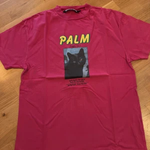 Palm Angels rosa t-shirt med kattmotiv - Palm Angels t-shirt i stark rosa med tryck av svart katt och texten 'PALM' i gult framtill. Oversized passform och klassisk rund hals. Tillverkad i 100% bomull för en skön känsla. Perfekt statement-plagg för dig som gillar streetwear och coola prints.