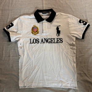 Vit Polo Ralph Lauren Los Angeles piké - Vit pikétröja från Polo Ralph Lauren med svart krage och detaljer. Broderad Los Angeles-text, stort polospelare-logo och emblem på bröstet samt siffra 3 på ärmen. Klassisk och sportig look, perfekt för dig som gillar streetstyle.