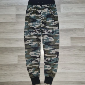 Camo mjukisbyxor med dragkedjor - Snygga mjukisbyxor (tunna) i camouflage-mönster med svarta muddar i midja och benslut. Byxorna har två fickor framtill med dragkedjor i guld och är perfekta för en avslappnad streetstil. Materialet är mjukt och bekvämt, passar perfekt till chill eller träning.