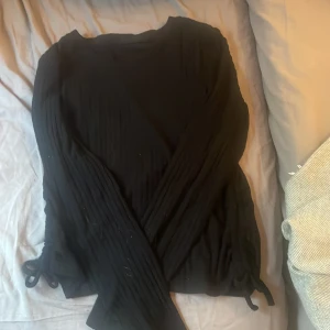 Svart ribbad långärmad topp Xoxo Friends - Svart långärmad topp från Xoxo Friends med ribbad struktur och knytband vid ärmsluten. Toppen har en enkel rund halsringning och är i ett mjukt, stretchigt material som sitter snyggt på kroppen.