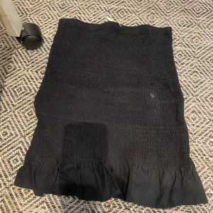 Svart volangkjol från Holly & Whyte - Svart kortkjol från Holly & Whyte med smockad midja och volangkant nertill. Kjolen är i mjuk bomull och har en enkel, stilren look som är lätt att matcha. Perfekt för dig som gillar en feminin och trendig stil.