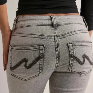 Grå bootcut jeans med broderade fickor - Snygga grå jeans med bootcut passform och låg midja. Jeansen har coola broderade detaljer på bakfickorna och klassisk femficksdesign. Perfekta för dig som vill ha en trendig och avslappnad look.