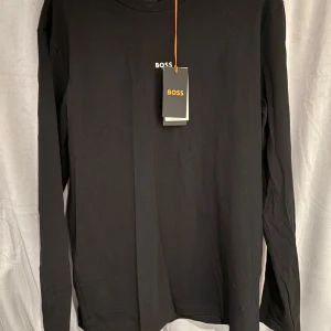 Svart långärmad tröja från BOSS - Svart långärmad tröja från BOSS med stilren design och liten vit logga på bröstet. Tillverkad i mjuk bomull som känns skön mot huden. Perfekt för dig som gillar en clean och klassisk look. Passar dig som vill ha en enkel men snygg tröja.