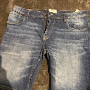 Blå jeansshorts från Produkt XL - Snygga blå jeansshorts från Produkt i storlek XL. Klassisk femficksmodell med gul kontrastsöm och knappstängning fram. Shortsens tvättade denim ger en cool och avslappnad vibe, perfekt för varma dagar. Märkespatch baktill och slitningar för extra stilpoäng.
