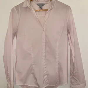 Ljusrosa skjorta med knappar - Säljer en stilren ljusrosa skjorta med klassisk krage och knäppning framtill. Skjortan har lång ärm och en figurnära passform som ger en snygg siluett. Perfekt att styla med jeans eller kostymbyxor för en clean look.