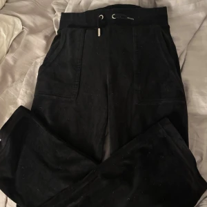 Svarta mjukisbyxor Juicy Couture XXS - Svarta mjukisbyxor från Juicy Couture i storlek XXS. Byxorna har dragsko i midjan, raka ben och stora fickor fram. Materialet är mjukt och skönt, perfekt för chill dagar. Snygga silverdetaljer på fickorna bak och klassisk Juicy Couture-logga.