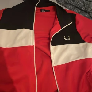 Röd och svart track jacket Fred Perry - Säljer en snygg track jacket från Fred Perry i storlek S. Jackan har breda block i rött, svart och vitt, med klassisk Fred Perry-logga på bröstet. Materialet är en mix av polyester och bomull, och jackan har dragkedja framtill. Perfekt för dig som gillar sportig stil. Pris kan diskuteras 