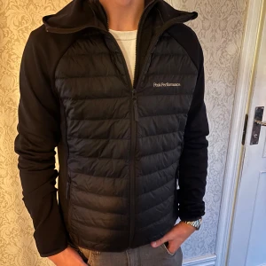 Svart hybridjacka Peak Performance L - Svart hybridjacka från Peak Performance i storlek L. Jackan har quiltad framsida och rygg, mjuka ärmar och huva. Stängs med dragkedja och har diskret logga på bröstet. Perfekt för kyliga dagar med sportig och clean look.