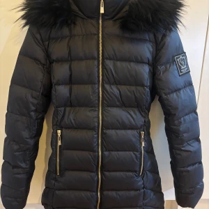 Svart pufferjacka från Chiara Forthi - Snygg svart pufferjacka från Chiara Forthi med fluffig fuskpäls runt huvan och guldiga dragkedjor. Jackan har quiltad design, lång arm och en patch med logga på ena ärmen. Perfekt för kalla dagar när du vill vara både varm och stilren.