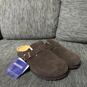 Bruna Birkenstock Boston mockatofflor - Säljer ett par bruna Birkenstock Boston i mocka med klassisk korksula och justerbart spänne i metall. Skorna har en stängd tå och öppen häl, perfekt för chill dagar. Mjuk innersula för extra komfort och stilren look.