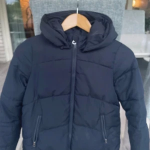 Svart pufferjacka från Vero Moda - Säljer en svart pufferjacka från Vero Moda med huva och dragkedja framtill. Jackan har två snedställda fickor med dragkedja och är perfekt för kyliga dagar. Enkel och stilren design som funkar till det mesta.