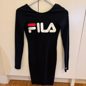 Svart tight klänning från FILA  - Svart, figurnära klänning från FILA i storlek XS. Klänningen har långa ärmar, rund halsringning, lite öppen rygg och stor vit FILA-logga framtill med röd detalj. Mjuk och stretchig bomullsblandning som sitter snyggt på kroppen.