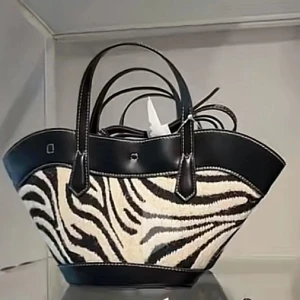 Zebramönstrad handväska med skinn - Cool handväska med svart-vitt zebramönster och svarta skinn-detaljer. Väskan har två handtag och en unik, bred form som sticker ut. Perfekt för dig som vill ha en statement-accessoar med trendig djurprint och snygga kontraster.