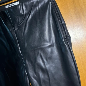 Svarta byxor i skinnimitation  - Byxor i skinnimitation från Indiska med rak passform och dragkedja i sidan. Ser skrynkliga ut på bild men det slätas ut när de är på. 