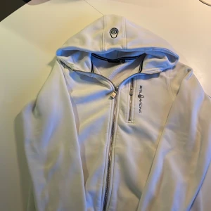 Vit hoodie från Sail Racing med dragkedja - Vit hoodie från Sail Racing med hel dragkedja och broderad logga på bröstet. Tröjan har huva, lång ärm och en bröstficka med dragkedja. Mjuk insida och sportig look, perfekt för dig som gillar stilrena och funktionella plagg. Storlek 160