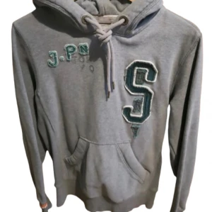 Superdry hoodie - Grå hoodie med broderade bokstäver och stor S på bröstet. Tröjan har huva med snörning och en klassisk känguruficka framtill. Perfekt för en avslappnad och sportig stil.