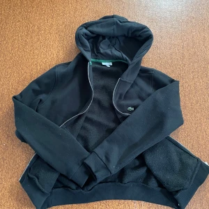 Svart hoodie från Lacoste med dragkedja - Svart hoodie från Lacoste med klassisk krokodillogga på bröstet. Tröjan har huva, långa ärmar och dragkedja framtill. Tillverkad i mjuk bomullsmix med ribbade muddar och nederkant. Perfekt för chill dagar och streetstyle.