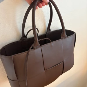 Brun handväska  - Snygg FEJK brun handväska i flätad design. Väskan är tillverkad i skinn och har två rejäla handtag samt en innerficka med dragkedja. Perfekt storlek för att få plats med allt du behöver och stilren look som lyfter din outfit. 
