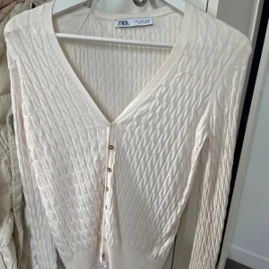 Beige kofta med knappar från Zara - Snygg beige kofta från Zara i storlek S med kabelstickat mönster och v-ringning. Koftan har långa ärmar och flera små, transparenta knappar framtill. Perfekt för lager-på-lager och chill stil. Materialet känns mjukt och är stickat.
