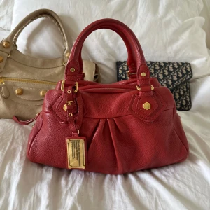 Röd Q Classic Baby Groove väska! - Snyggaste röda handväskan i äkta läder från Marc by Marc Jacobs. Den ikoniska modellen Q Classic Baby Groove! En populär och eftertraktad vintagemodell med gulddetaljer och dubbla handtag. Väskan är i nyskick. Lädret är helt utan repor eller hål.   Det här är en tidlös modell som inte längre finns att köpa, vilket gör den extra eftertraktad. Perfekt för dig som vill ha en stilren tidlös väska med riktigt bra andrahandsvärde. Passar lika bra till vardag som till fest.  Mått: 34x18x12 cm 🤍