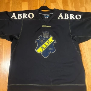 AIK hockeytröja från 2012 - Säljer en svart AIK hockeytröja från Bauer i storlek XL. Tröjan har AIK:s klubbmärke på bröstet, stora vita siffror 84 på ryggen och gula detaljer på ärmarna. Broderad text Åbro på axlarna.