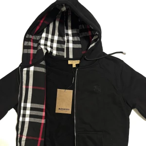 Svart zip hoodie från Burberry  - Svart vindjacka från Burberry med klassiskt rutigt foder i huvan. Jackan har dragkedja framtill, snörning i huvan och broderad logga på bröstet. Tillverkad i bomull och polyester för en skön känsla. Perfekt för dig som vill ha en stilren och tidlös look. Perfekt för de kommande kyliga dagar. Skickas inom 24 timmar. 