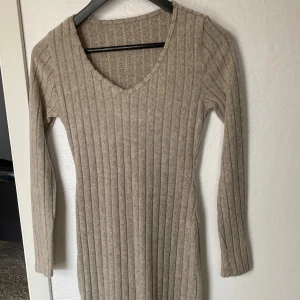 Beige ribbad stickad klänning S - Ribbstickad beige klänning i mjuk ull med lång ärm och v-ringning. Modellen är kroppsnära och har en klassisk, enkel design som passar perfekt till kyligare dagar. Klänningen är midilång och har en stilren look.