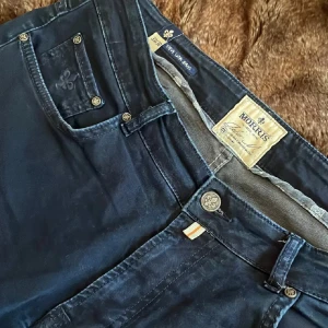 Morris blå byxor i bomull - Snygga blå byxor från Morris i mjuk bomull med klassisk femficksdesign och diskreta logodetaljer på knappar och ficka. Byxorna har normal passform och passar perfekt till en stilren look. Nere vid fotbenet på bak av jeansen så är det lite av en ljusare blå! Pågrund av lite slitning 