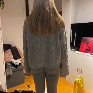 Grå kabelstickad kofta med krage - Mysig grå kabelstickad kofta med bred krage och v-ringad öppning framtill. Tröjan har långärmade, lite loose passform och ribbade muddar vid ärmslut och nederkant. Perfekt för chill dagar och snygg att styla med jeans eller mjukisbyxor.