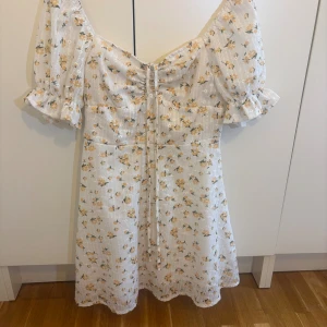 Vit blommig kortklänning från SHEIN - Supersöt vit kortklänning från SHEIN med gula blommor, puffärmar och snörning framtill. Klänningen har volangdetaljer vid ärmslut och en romantisk v-ringning. Perfekt för sommaren och vårens alla tillfällen!