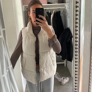 Beige quiltad dunväst med dragkedja - Säljer en beige quiltad dunväst med hög krage och dragkedja framtill. Västen har en loose passform och är perfekt att slänga över en hoodie eller långärmad tröja. Bra skick. Passar från s-m 