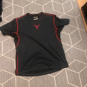 Svart Bauer t-shirt med röda detaljer - Svart kortärmad t-shirt från Bauer med röda sömmar och logga på bröstet. Modellen är sportig och har en bekväm passform, perfekt för dig som gillar en aktiv stil. Materialet är syntet och andas bra under träning. Storlek barn xl