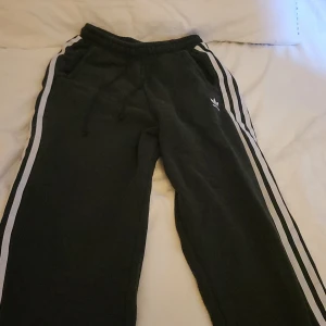 Svarta Adidas mjukisbyxor  - Svarta mjukisbyxor från Adidas med klassiska vita ränder längs sidorna och broderad logga på låret. Byxorna har resår och snörning i midjan samt bekväma sidofickor. Perfekta för chill eller sportig stil.