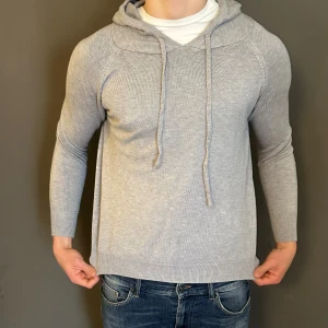 Ljusgrå stickad hoodie med snörning - Snygg ljusgrå stickad hoodie med huva och snörning framtill. Tröjan har långärmade ärmar och en mjuk, ribbad kant vid ärmslut och nederkant. Perfekt för dig som gillar en chill och enkel stil. Passar till jeans och sneakers.