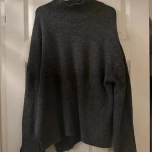 Mörkgrå stickad polotröja - Mysig mörkgrå stickad polotröja med oversized passform och långa ärmar. Tröjan har en hög polokrage och ribbade muddar vid ärmslut och nederkant. Perfekt för kyliga dagar och enkel att styla med jeans eller kjol.