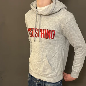 Grå hoodie Moschino MTV - Snygg grå hoodie från Moschino med MTV-logga i rött och vitt broderat framtill. Tröjan har huva med snörning, magficka och långa ärmar. Perfekt för dig som gillar streetwear och vill sticka ut med ett coolt tryck.