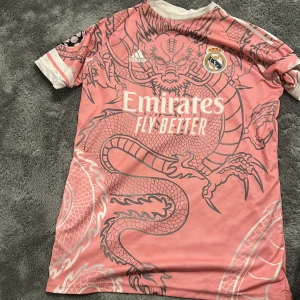 Rosa Real Madrid fotbollströja Adidas L - Säljer en rosa Real Madrid fotbollströja från Adidas med drakmönster och vita detaljer. Tröjan har korta ärmar, klubbmärke på bröstet, sponsortryck och nummer 15 med namnet Arda Güler på ryggen. Materialet är lätt och andas, perfekt för match eller träning.