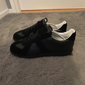 Maison Margiela svarta sneakers - Svarta sneakers från Maison Margiela med klassisk låg siluett och rund tå. Skorna har svarta snören, vit insida och är tillverkade i en mix av skinn och mocka. Kommer med originalkartong och dustbag. Perfekta för dig som gillar stilrena och exklusiva sneakers.