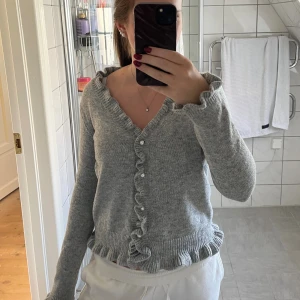 Grå stickad kofta med volang - Superfin grå stickad kofta med volangdetaljer längs knäppningen och nederkanten. Koftan har pärlknappar och en v-ringad hals som kan bäras offshoulder. Säljer då den inte kommer till användning🩶strl S