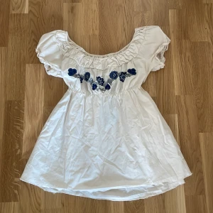 Odd Molly Blus - Supersöt vit blus ifrån Odd Molly med blått broderi framtill och baktill. Blusen har puffärmar, volang vid halsringningen och ett knytband i ryggen. Tidigare köpt ifrån Sellpy, oanvänd.