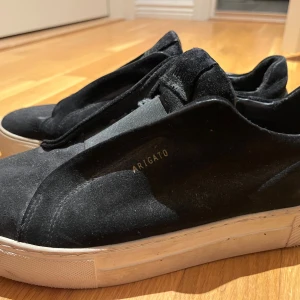 Svarta sneakers från Axel Arigato - Svarta sneakers från Axel Arigato med stilren design och vit platt sula. Ovandelen är i mjuk mocka och har elastiska paneler istället för klassisk snörning, vilket ger en clean look. Perfekta för dig som gillar minimalistisk stil och vill ha något som sticker ut lite. Man st