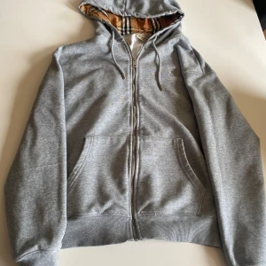 Grå Burberry hoodie med dragkedja - Säljer en grå hoodie från Burberry med klassiskt rutigt foder i huvan och broderad logga på bröstet. Tröjan har dragkedja, snörning i huvan och känguruficka. Perfekt för dig som vill ha en stilren och lyxig look.